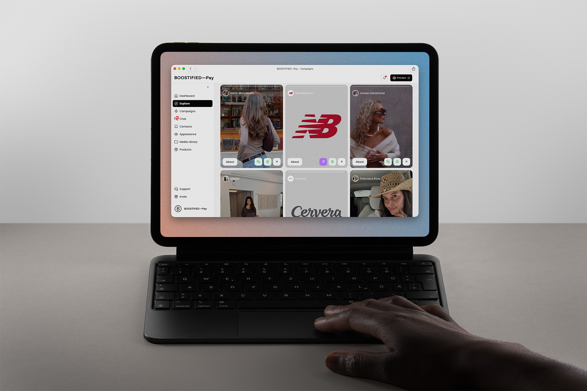 A tablet on a keyboard case showing the BOOSTIFIED—Pay creator explore view, with a hand resting on the trackpad