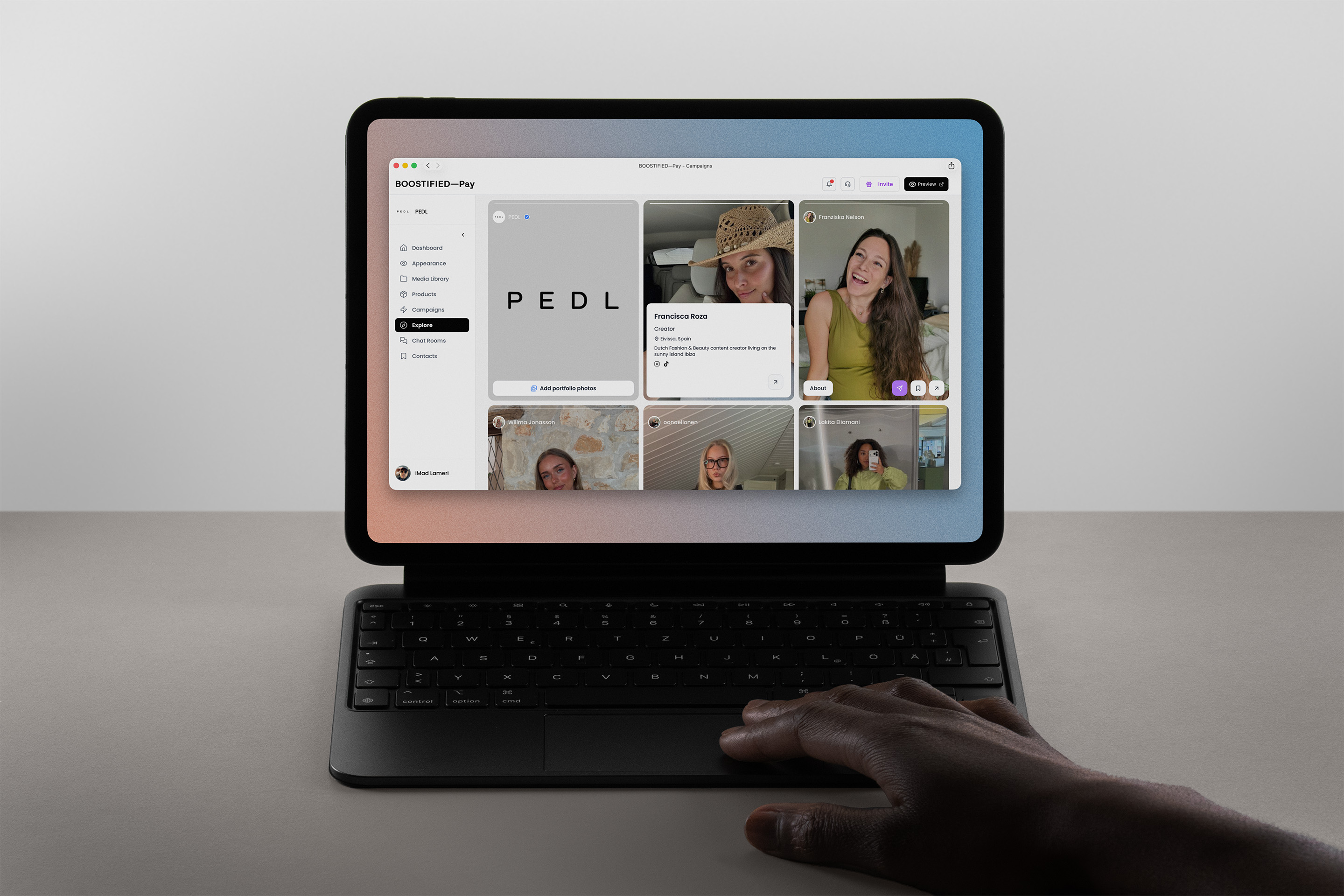 A tablet on a keyboard case showing the BOOSTIFIED—Pay creator explore view, with a hand resting on the trackpad