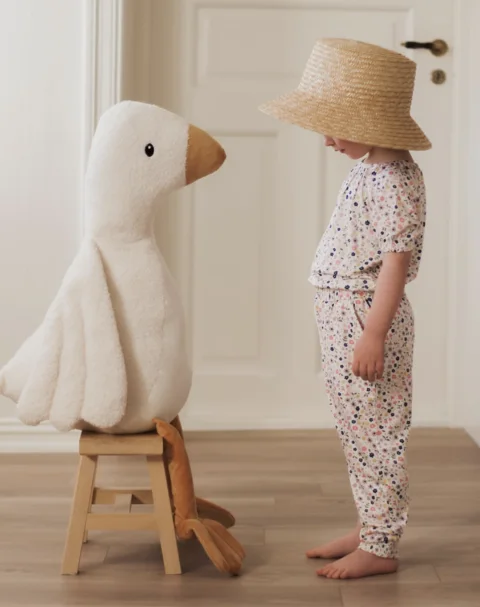 Child in floral clothing and a straw hat beside a plush goose